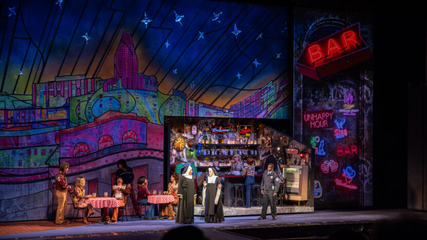 A colorful stage set inspired by Sister Act features actors in costume inside and around a neon-lit diner, with a vibrant cityscape backdrop. Some sit at checkered tables, while others stand in conversation. The audience is visible in the foreground.