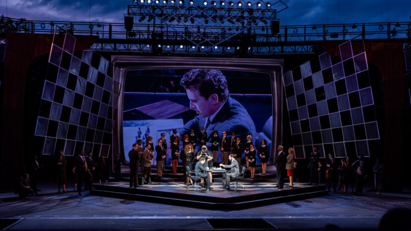 A dramatic stage scene features actors around a table, surrounded by people in formal attire, with a giant screen backdrop showing a close-up of a man playing chess, and an elaborate lighting rig overhead.