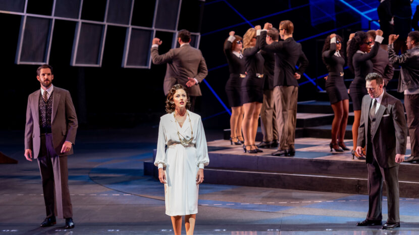 A woman in a light dress stands center stage like a chess queen, flanked by two men in suits, as a group in business attire with raised arms stand on tiered steps in the background, all under dramatic stage lighting.