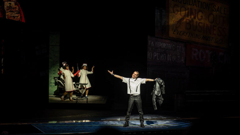 A man stands center stage with arms outstretched, holding a black jacket under a spotlight, evoking a scene from Little Shop of Horrors. In the background, two women in white stand near a green-tiled wall beside liquidation sale signs.