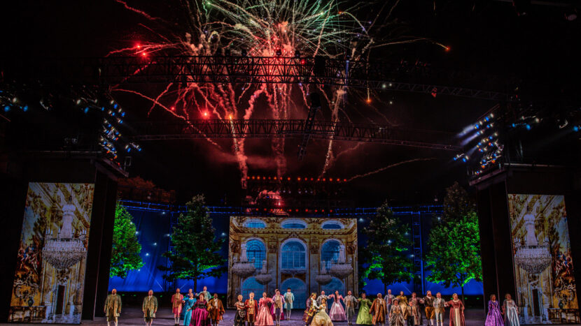 Performers in historical costumes stand on a grand outdoor stage with elaborate scenery, bringing Beauty and the Beast to life as colorful fireworks burst in the night sky above, creating a festive atmosphere.