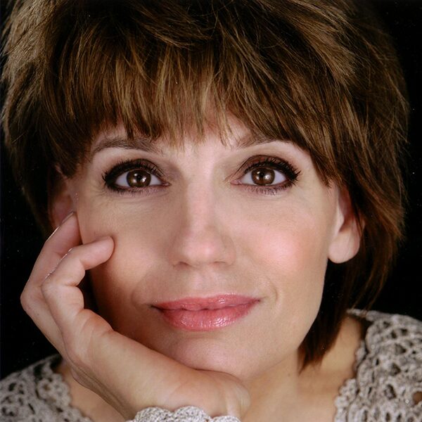 A woman with short brown hair and brown eyes gazes at the camera, resting her face on her hand. She is wearing a light-colored, textured top and has a calm, gentle expression.
