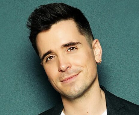 A man with short dark hair, wearing a black blazer, white shirt, and a small hoop earring, smiles slightly while standing against a teal background.