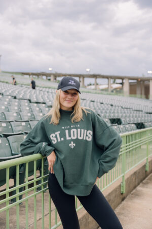 Wearing a dark green Meet Me Mockneck and a black cap, a person stands beside green stadium seats, smiling with one arm resting on the railing. The sky is cloudy and the stadium is mostly empty.