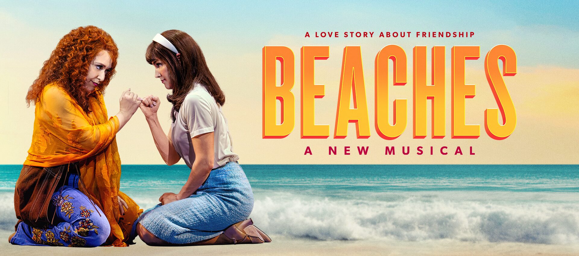 Two women kneel on a beach, holding hands and facing each other. The text above them reads, “A love story about friendship. BEACHES. A New Musical.” Ocean waves and a pastel sky are in the background.
