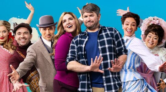 A joyful group of people in colorful, old-fashioned costumes stands in front of a bright sky backdrop, smiling and posing playfully with enthusiastic expressions and open arms.