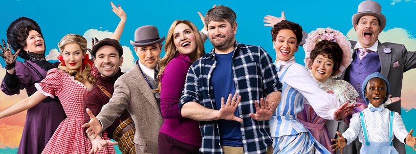 A joyful group of people in colorful, old-fashioned costumes stands in front of a bright sky backdrop, smiling and posing playfully with enthusiastic expressions and open arms.