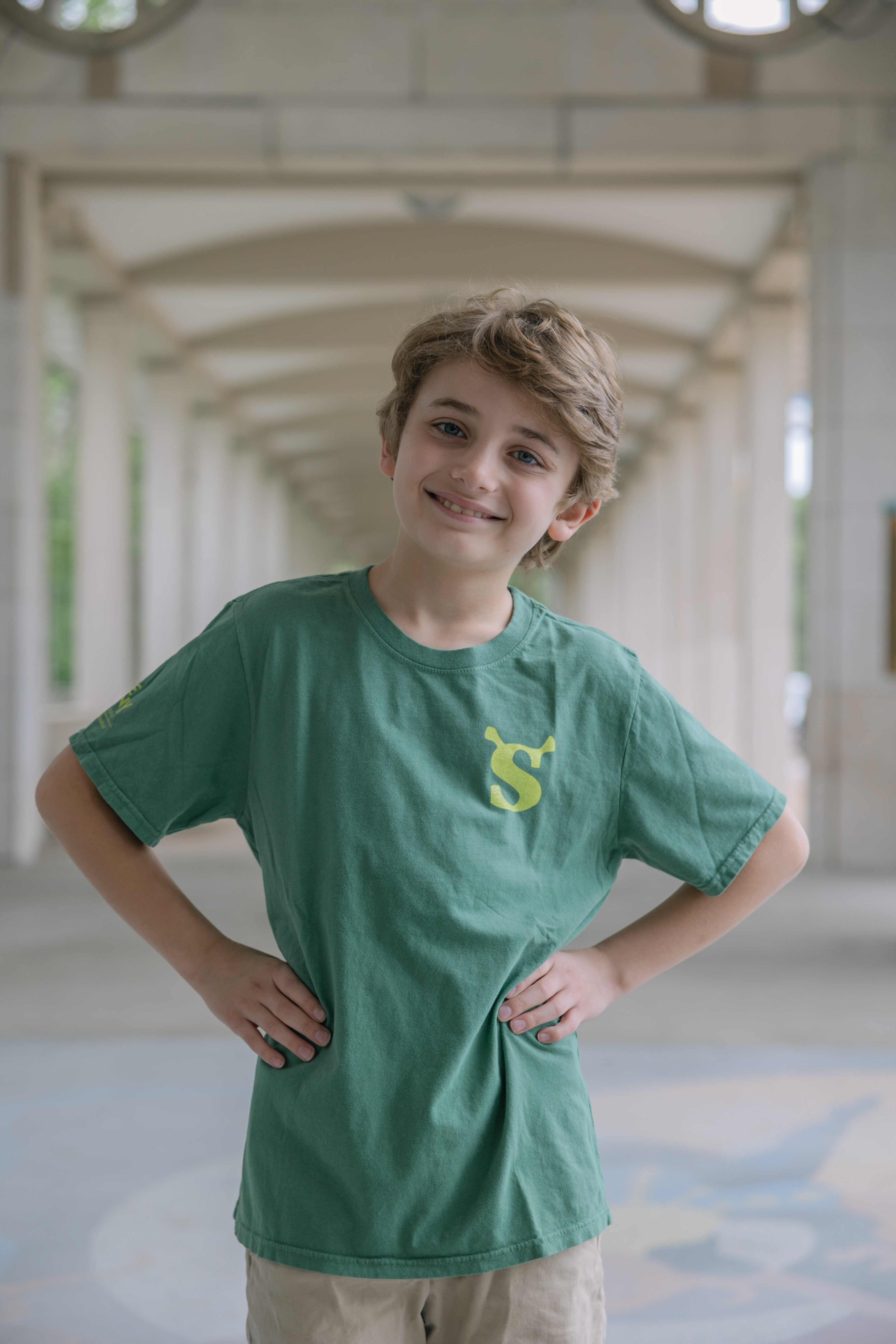 A young boy with light brown hair stands smiling with his hands on his hips, wearing the Shrek Show Shirt (Youth Sizes) and beige shorts in a bright, covered outdoor walkway with arches in the background.