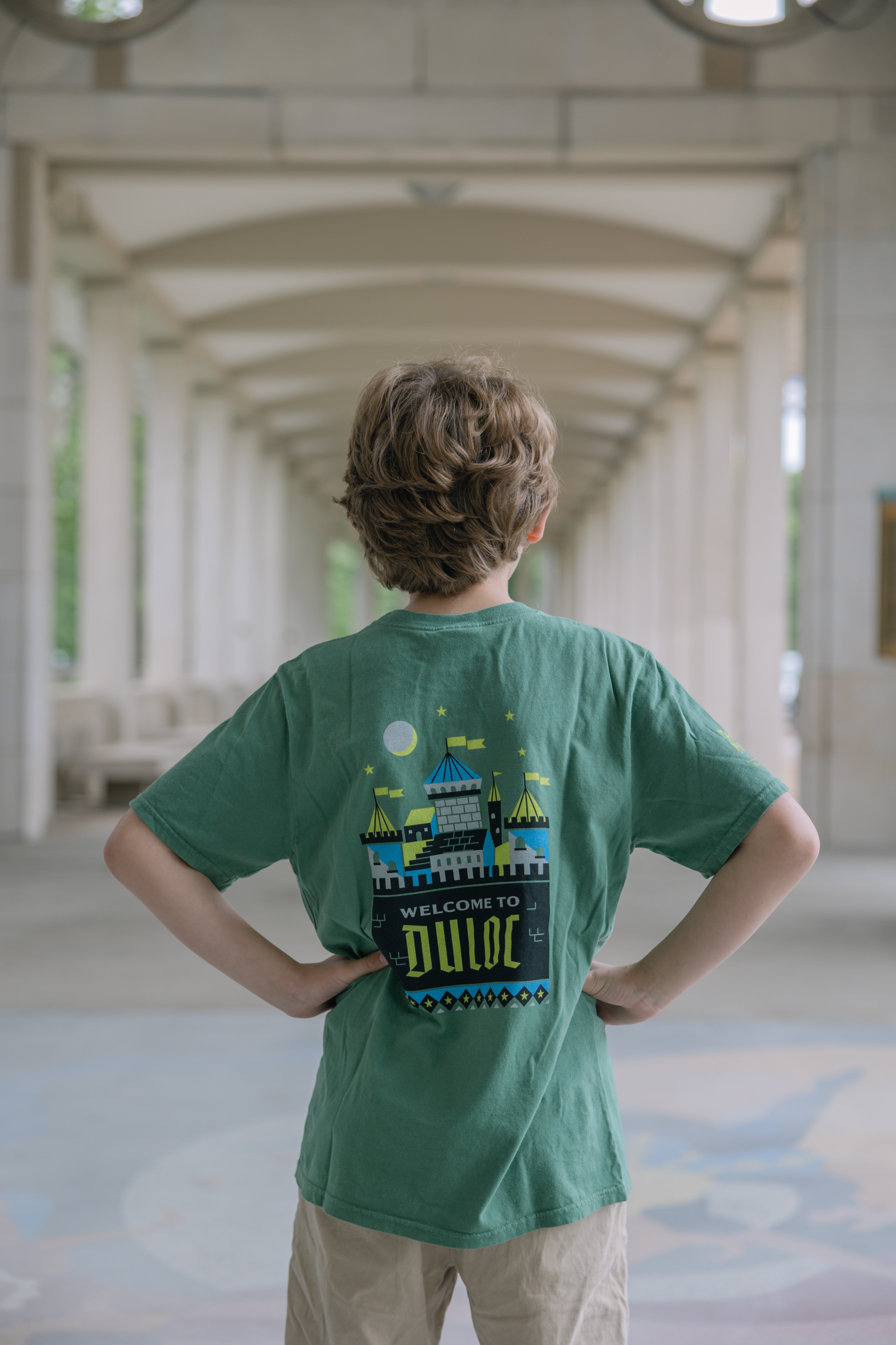 A child with short, light brown hair stands hands-on-hips, facing away in a green Shrek Show Shirt (Youth Sizes) and beige shorts, under a covered walkway with repeating arches.