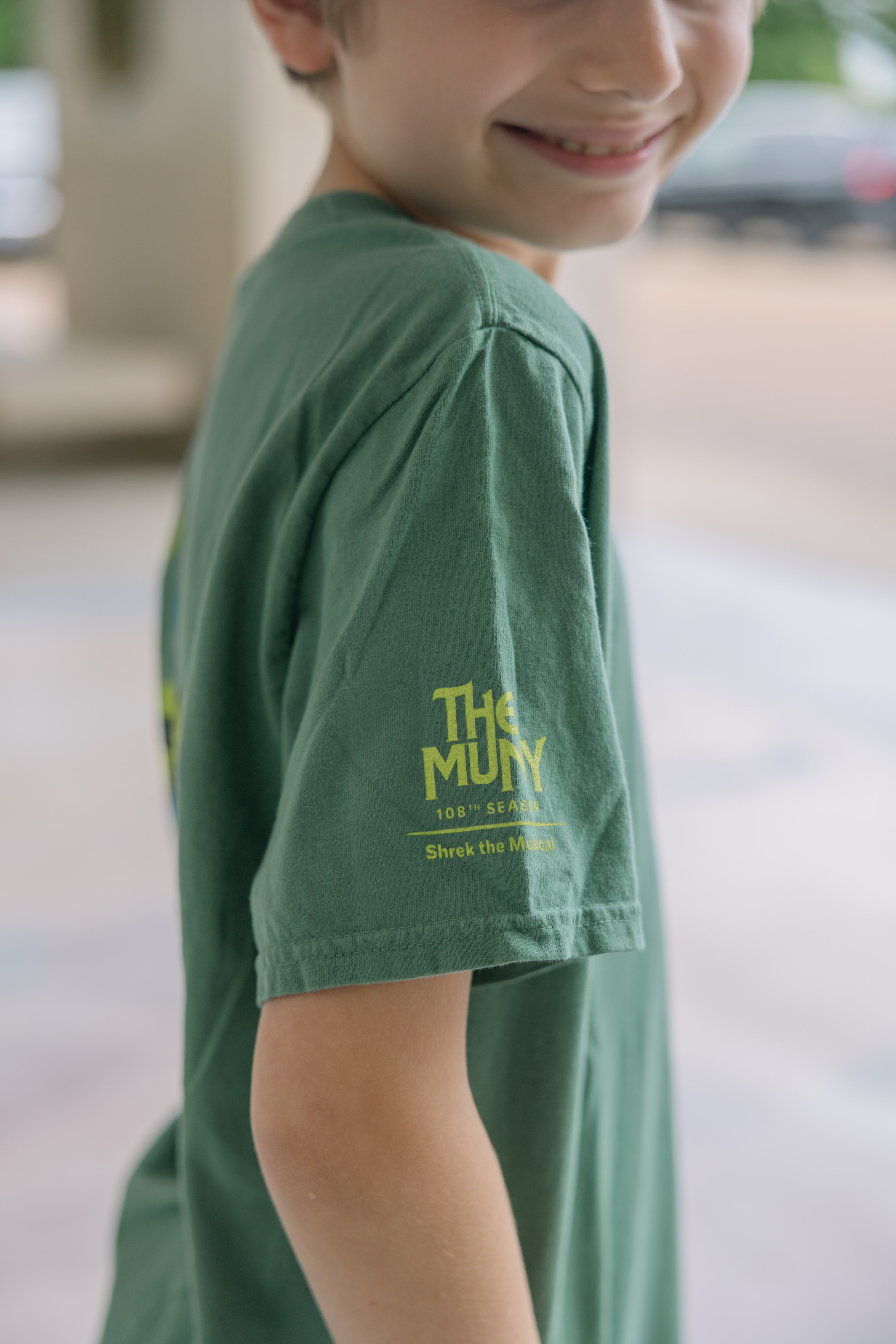 A smiling child wears the Shrek Show Shirt (Youth Sizes), a green T-shirt with yellow sleeve text, "The Muny 105th Season Shrek the Musical," against a softly blurred background.