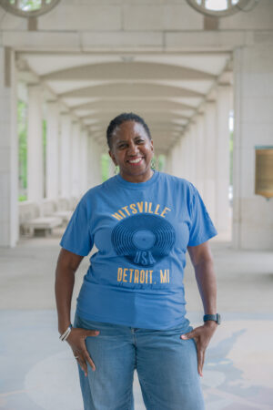 A woman stands smiling in a sunlit, arched walkway, wearing the blue "Ain't Too Proud Show Shirt" with “Hitsville USA Detroit, MI” on it and jeans. Light columns and trees appear through the arches in the background.