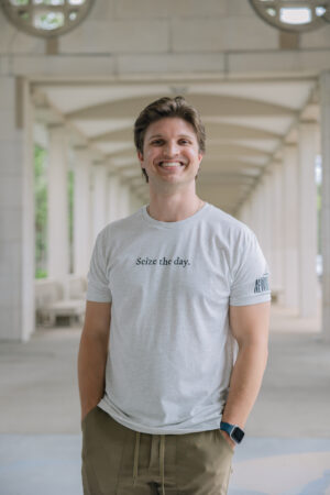 A smiling man stands under an arched walkway, hands in pockets, wearing a light gray Newsies Show Shirt, khaki pants, and a smartwatch. Columns and soft natural light appear in the background.