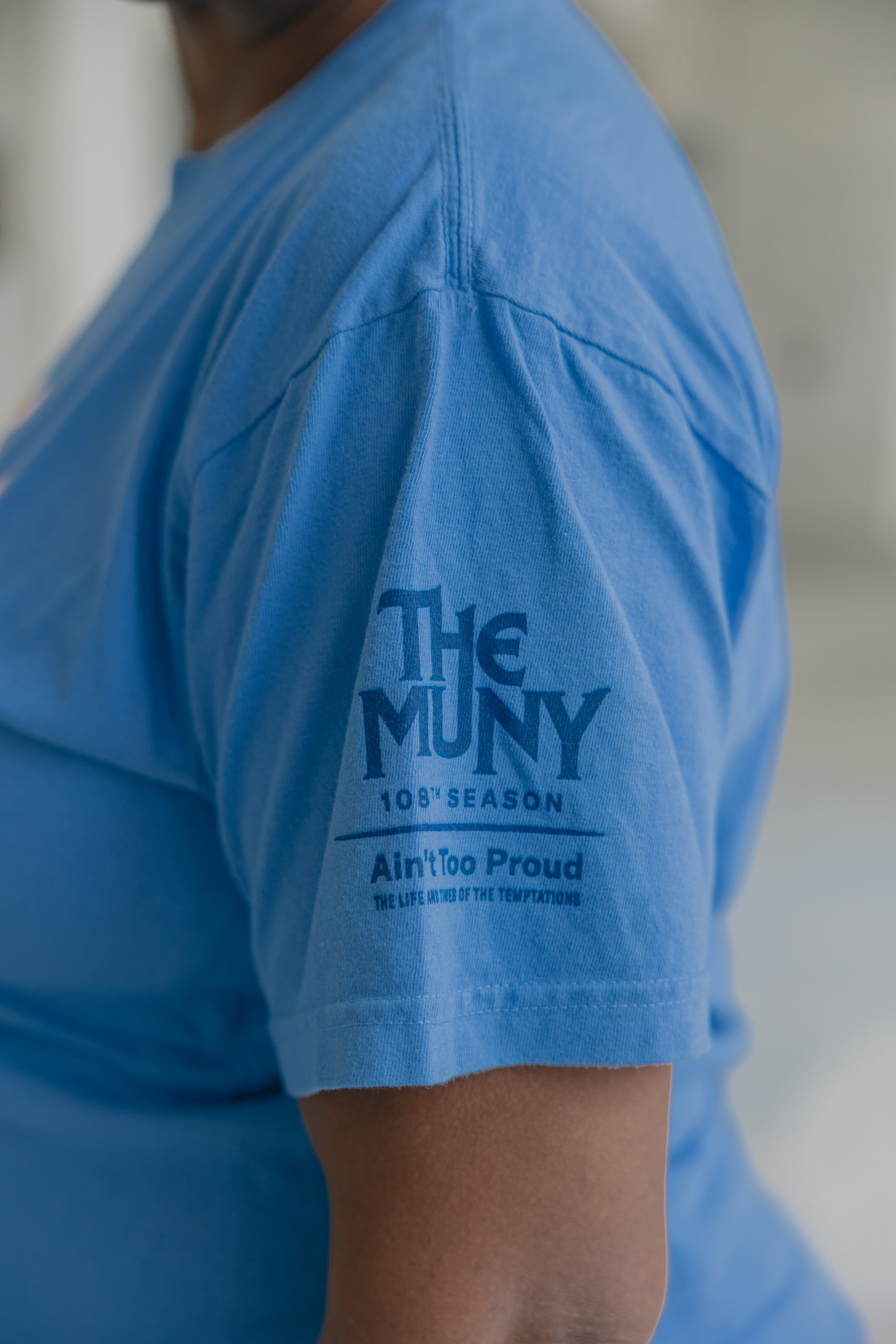 A person wearing the Ain't Too Proud Show Shirt, a blue T-shirt with "THE MUNY 105th SEASON Ain't Too Proud THE LIFE AND TIMES OF THE TEMPTATIONS" printed on the sleeve in dark blue text.
