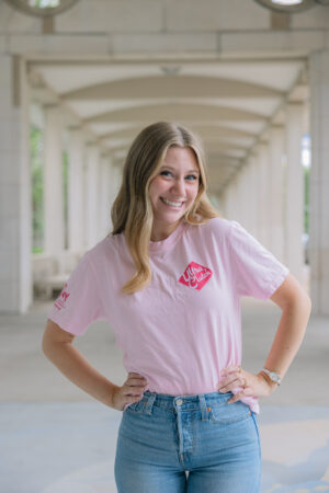 A smiling woman with long blonde hair stands under arched beams, wearing the Hairspray Show Shirt in light pink and blue jeans, hands on her hips.