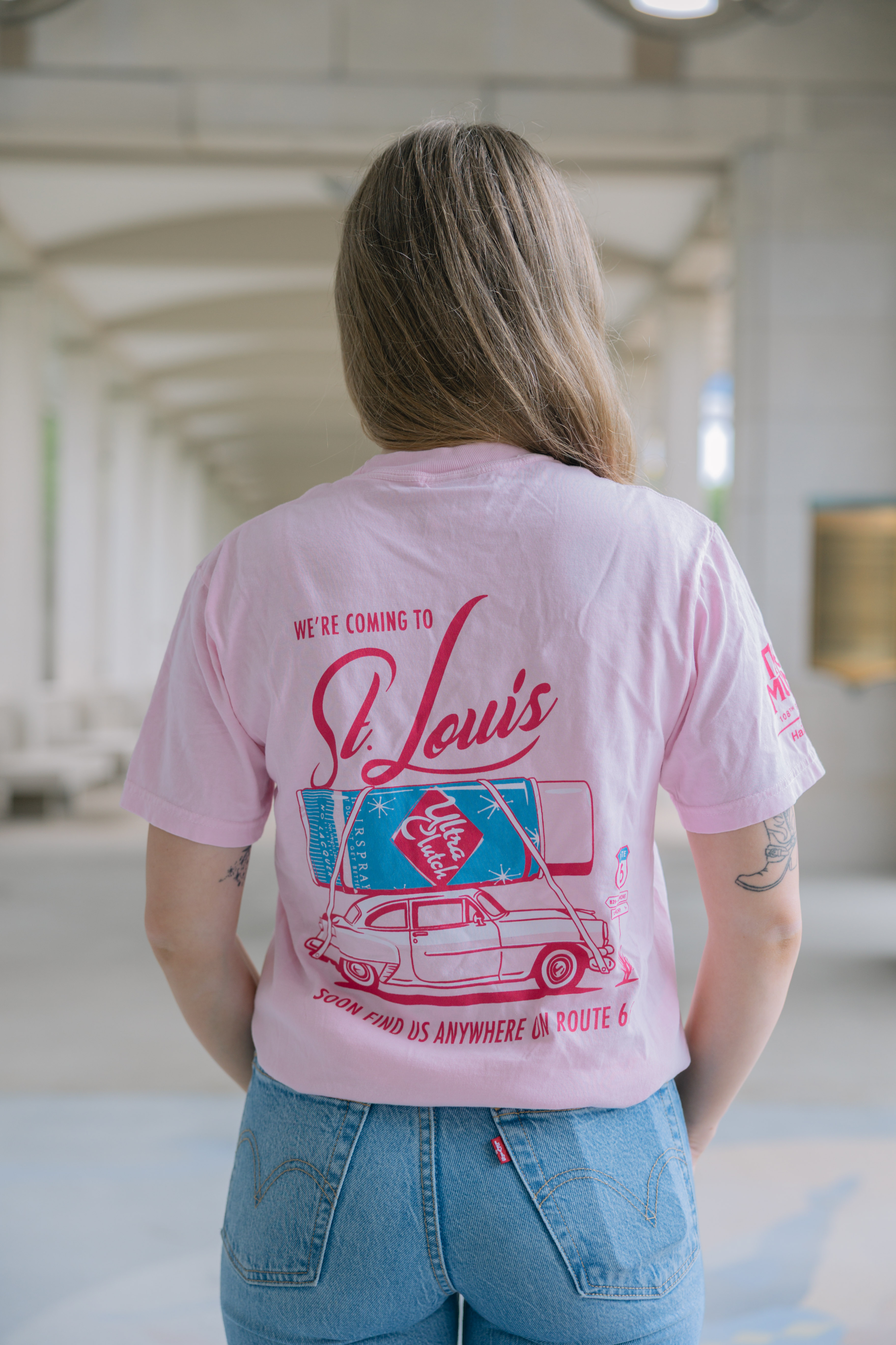 Wearing the Hairspray Show Shirt—a light pink tee with a retro "We're coming to St. Louis" car graphic on the back—paired with light blue jeans, this person stands in an outdoor corridor.