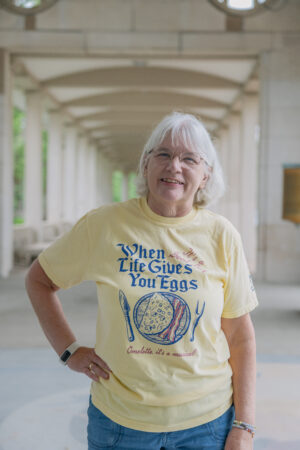 An older woman with white hair and glasses smiles outdoors, wearing the Something Rotten! Show Shirt—a yellow tee that says, “When life gives you eggs, omelette it’s a musical!” with egg, fork, and spoon illustrations.