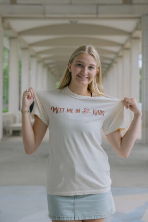 A young woman with long blonde hair smiles under an arched walkway, holding out the sides of her Meet Me In St.Louis Show Shirt with “MEET ME IN ST. LOUIS” in red letters, paired with a denim skirt.