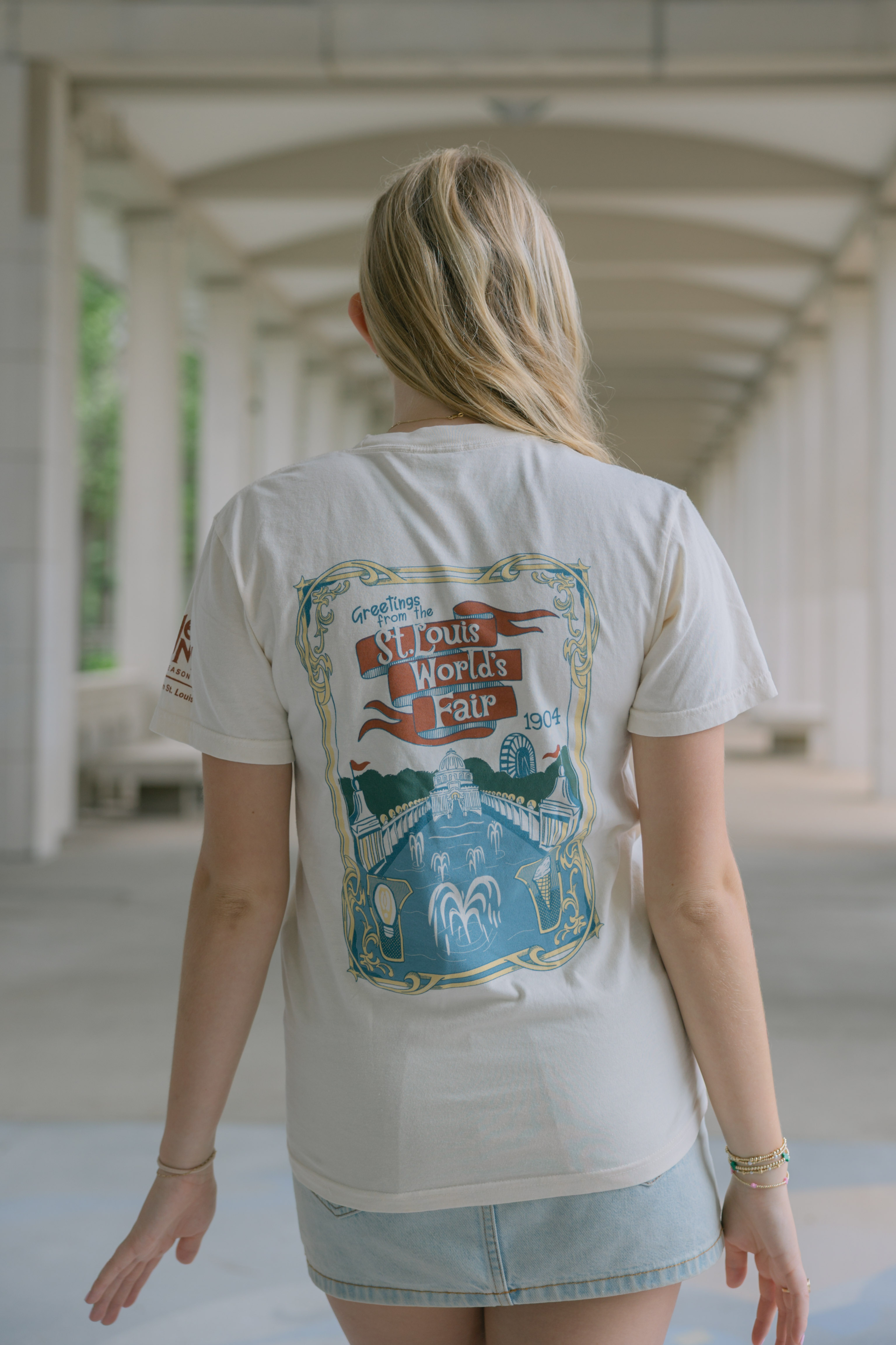 A person with long blonde hair, seen from behind, wears the Meet Me In St.Louis Show Shirt—a white tee with a colorful 1904 World's Fair design—while standing in a corridor with white columns and a geometric ceiling.