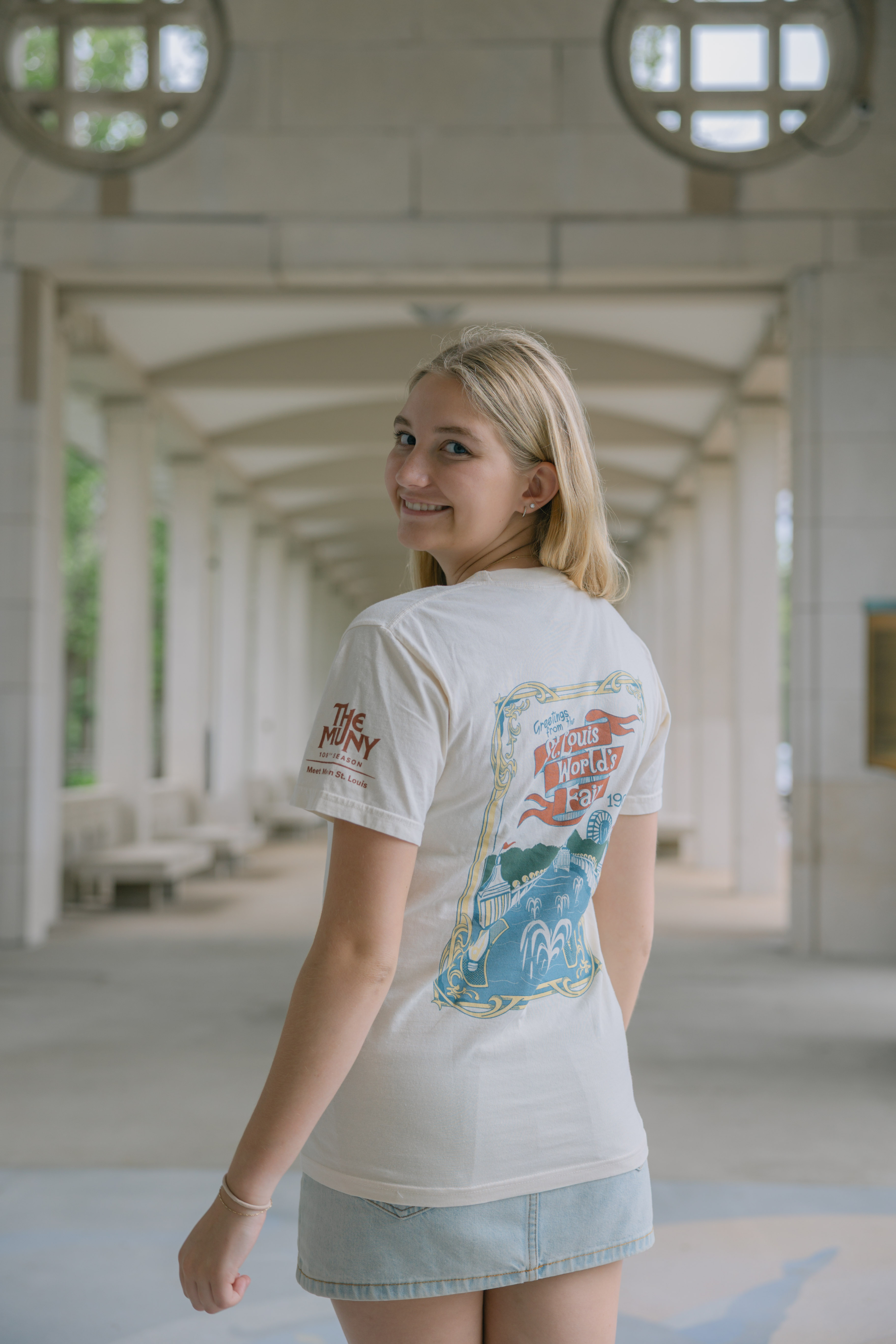 A young woman with blonde hair wears the Meet Me In St.Louis Show Shirt—a white tee with colorful graphics on the back—paired with a denim skirt, smiling over her shoulder while standing outdoors near arched walkways and round windows.