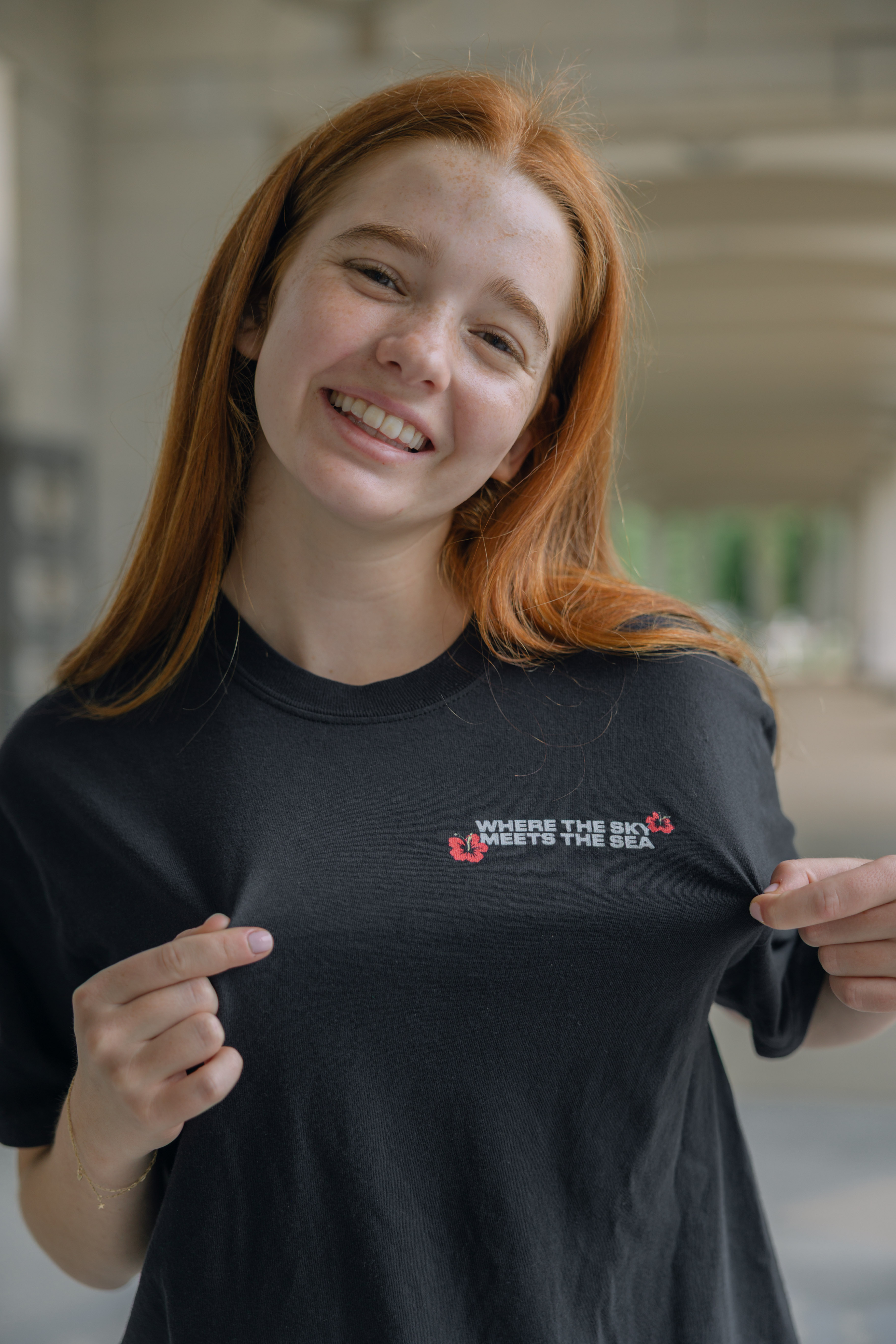 A smiling young woman with long red hair stands outdoors in a bright, covered walkway, holding out her black South Pacific Show Shirt featuring the text "WHERE THE SKY MEETS THE SEA.