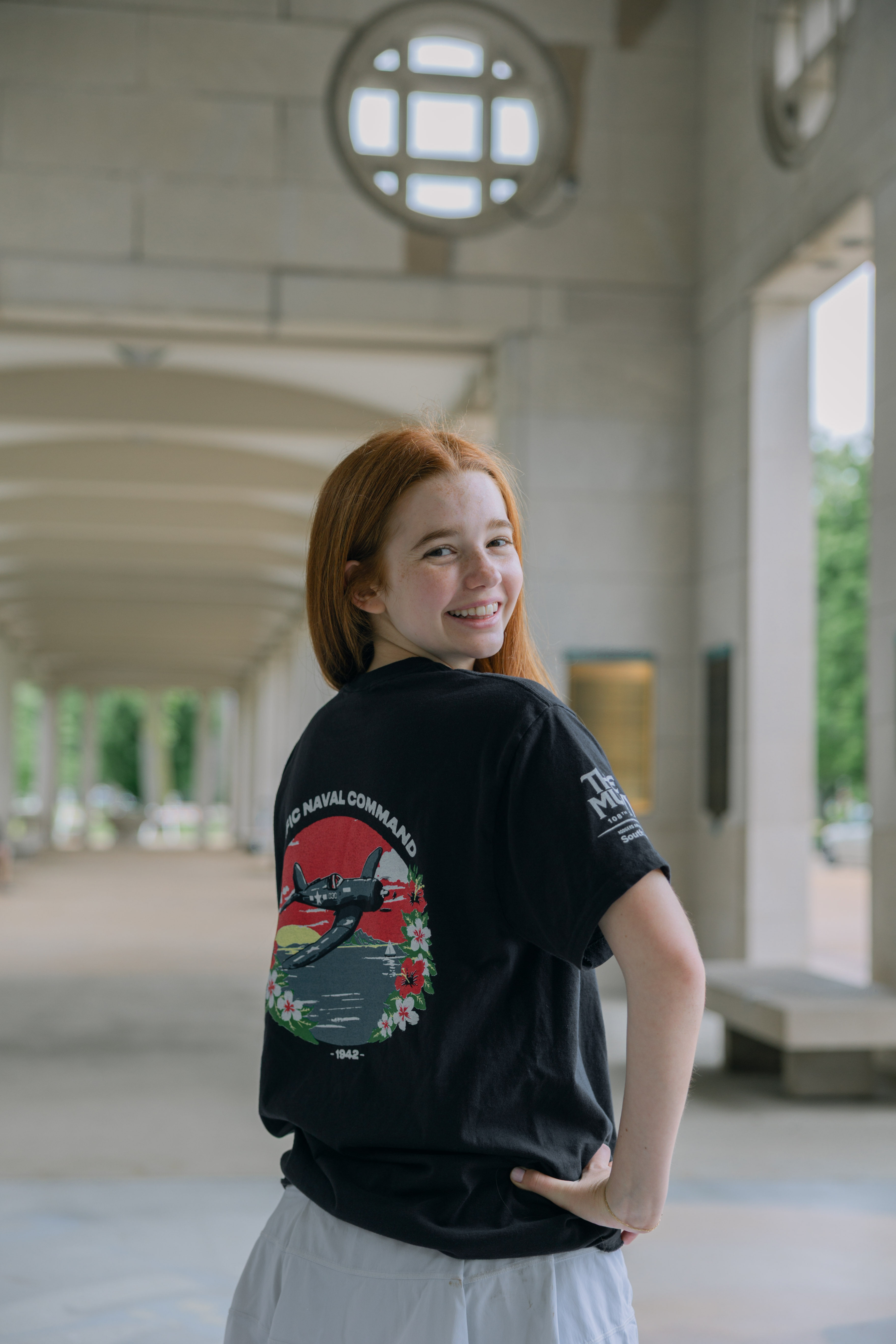 A young woman with long red hair smiles over her shoulder, wearing the South Pacific Show Shirt featuring a colorful back graphic and a white skirt, as she stands in a sunlit, arched outdoor corridor.