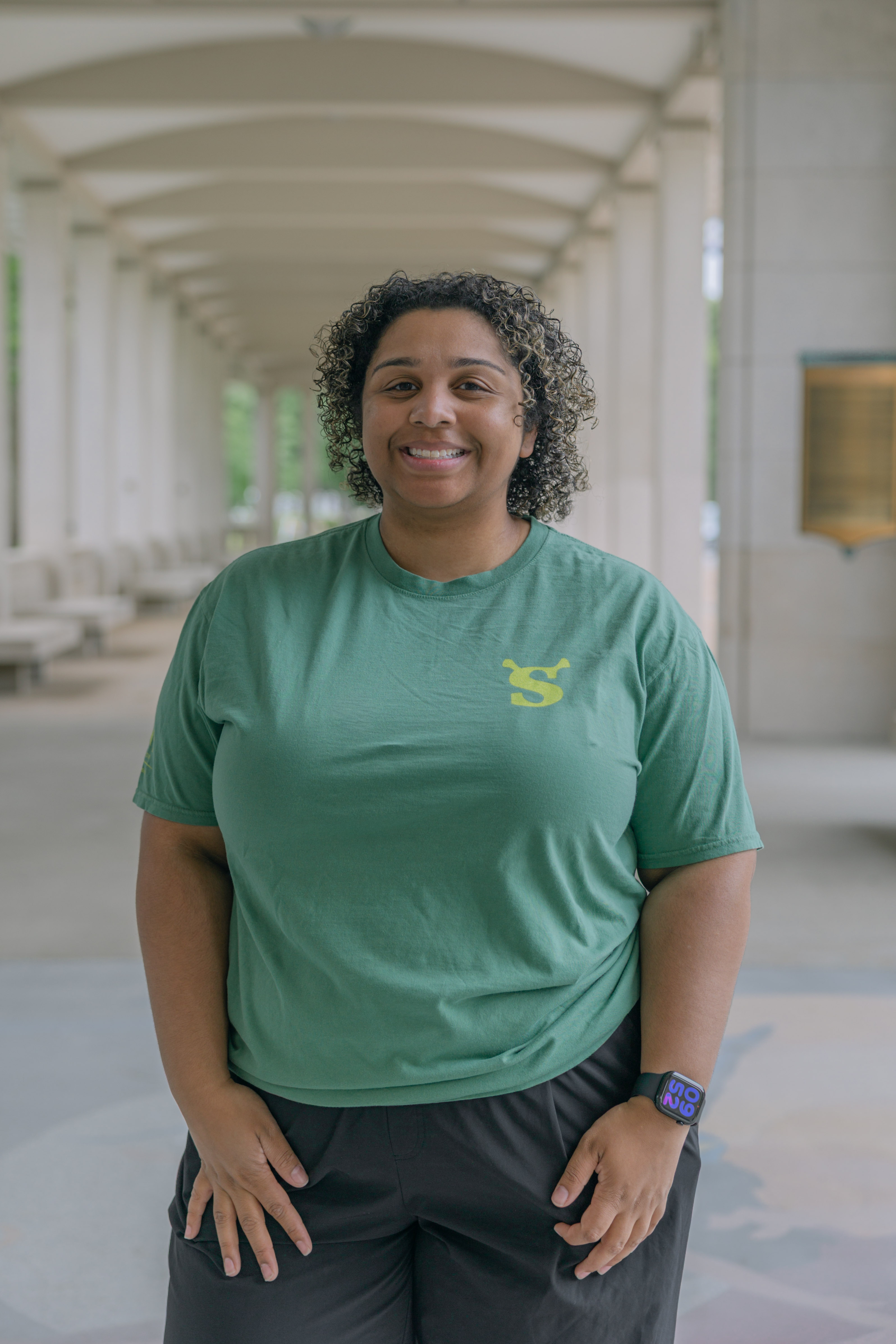 A person with short curly hair smiles in a covered outdoor walkway, wearing the Shrek Show Shirt (Adult Sizes). The green shirt features a yellow logo, and they also sport a blue watch. Columns and benches appear in the background.