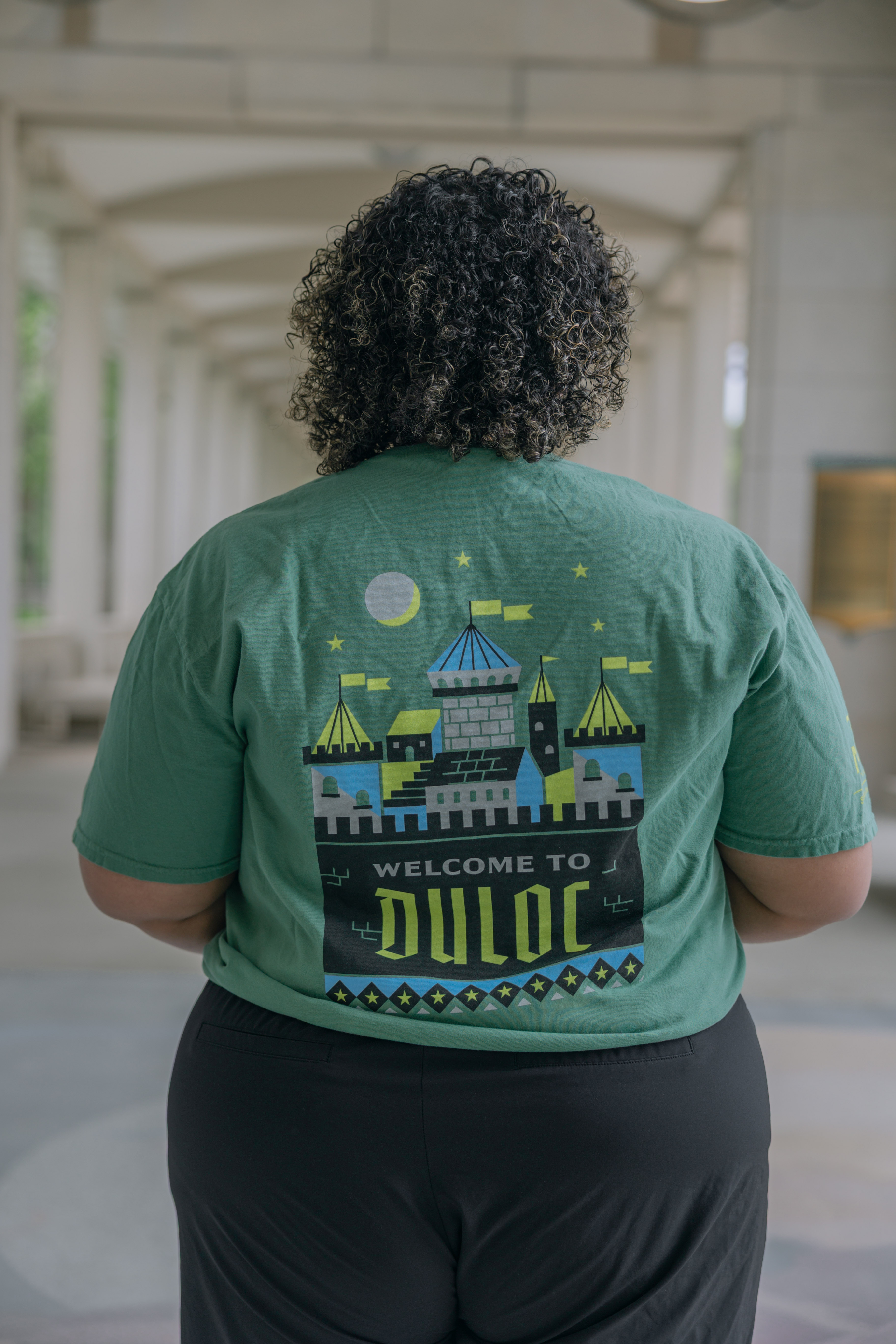 Seen from behind, a person with curly hair wears the Shrek Show Shirt (Adult Sizes), a green shirt with an illustrated castle, stars, and "WELCOME TO DULOC" text. The background has a covered walkway with columns.