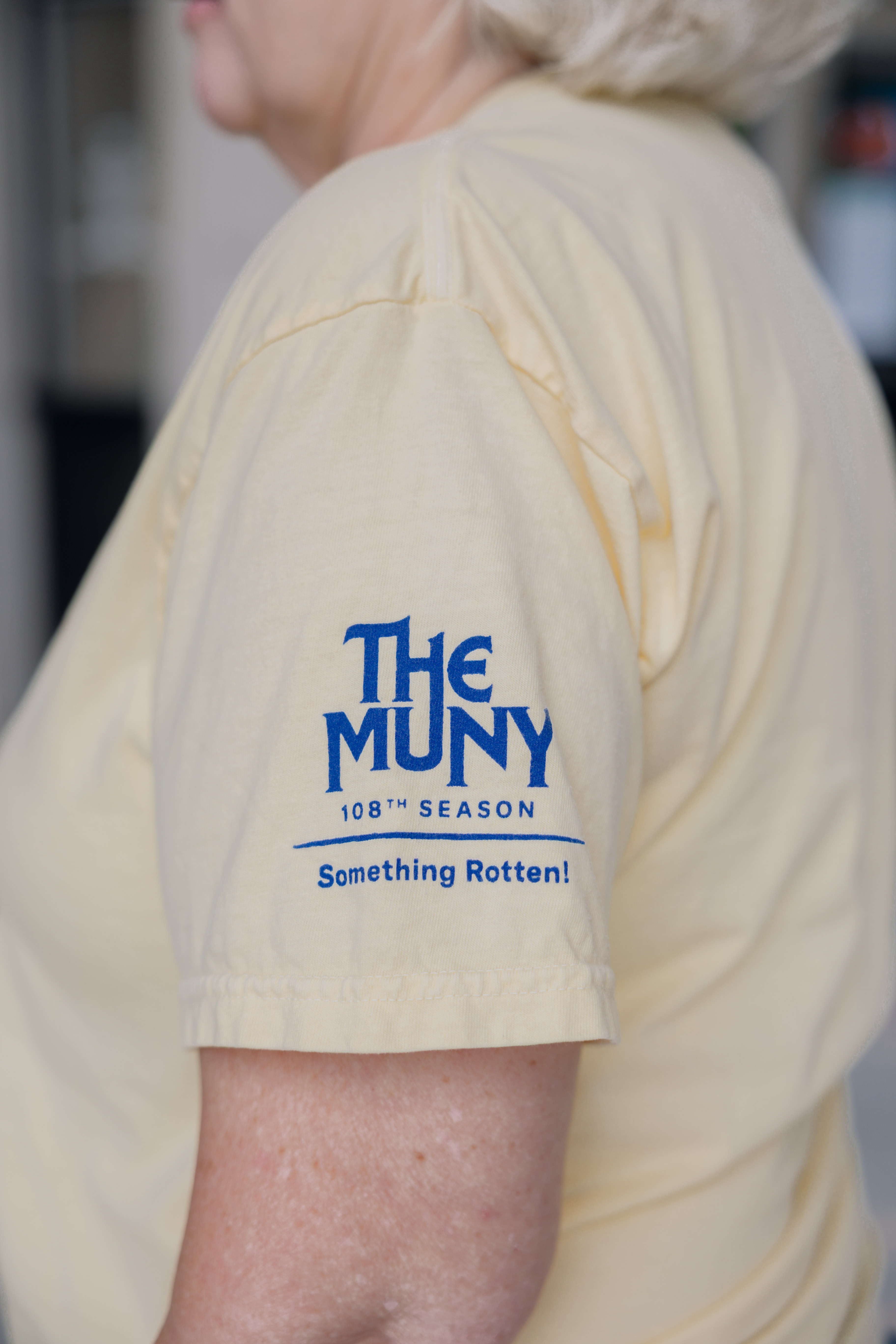 A person is wearing the Something Rotten! Show Shirt, a pale yellow tee featuring blue “The Muny 108th Season Something Rotten!” text on the sleeve. Only the shoulder, arm, and part of their face are visible.