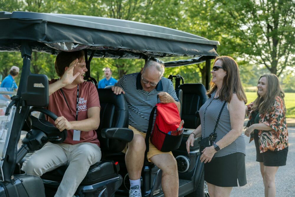 A group of adults gather around a parked golf cart outdoors. One person waves from the drivers seat, while another with a red bag prepares to sit inside. Two women stand nearby, smiling and chatting, with trees in the background.