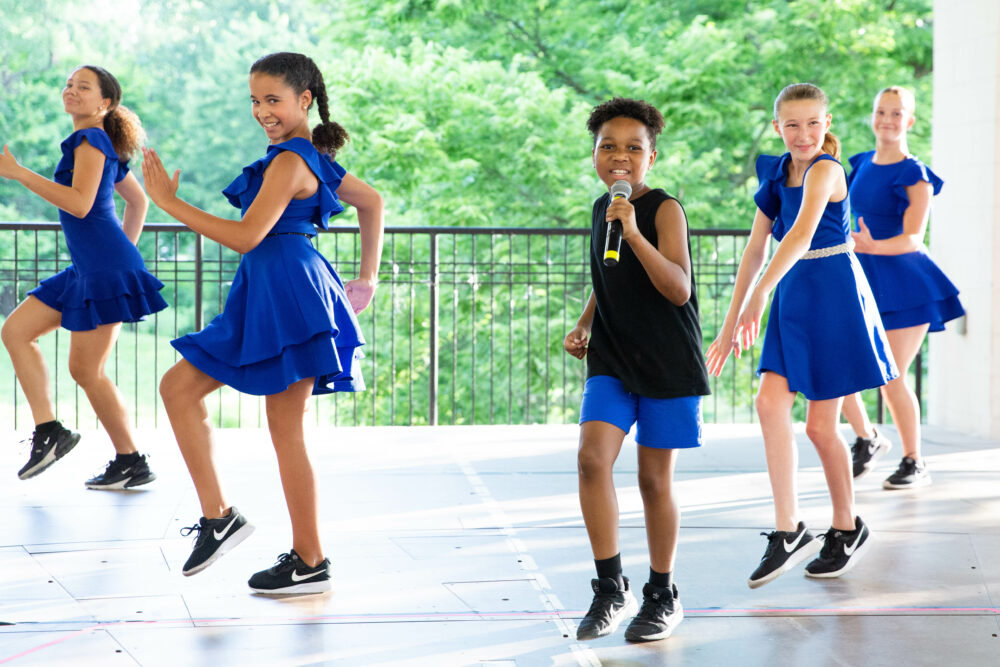 Five children from Muny Kids & Teens perform on stage; four girls in matching blue dresses dance, while a boy in a black shirt and blue shorts sings into a microphone. Green trees are visible in the background.