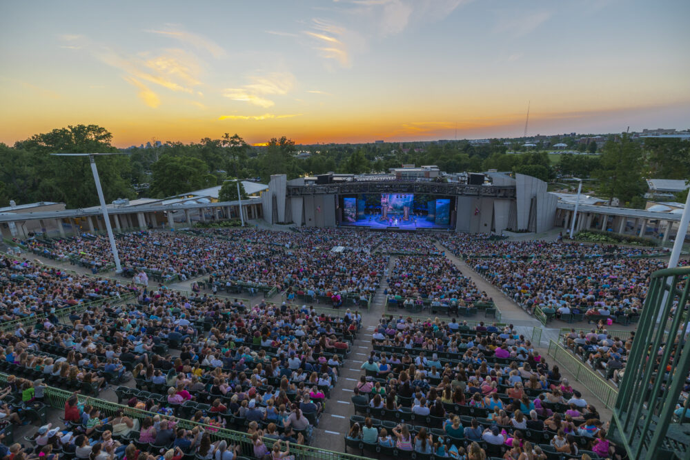 An outdoor amphitheater filled with people enjoying a live performance at sunset, the stage brightly lit against trees and cityscape—experience memorable nights and ticket savings under the open sky.