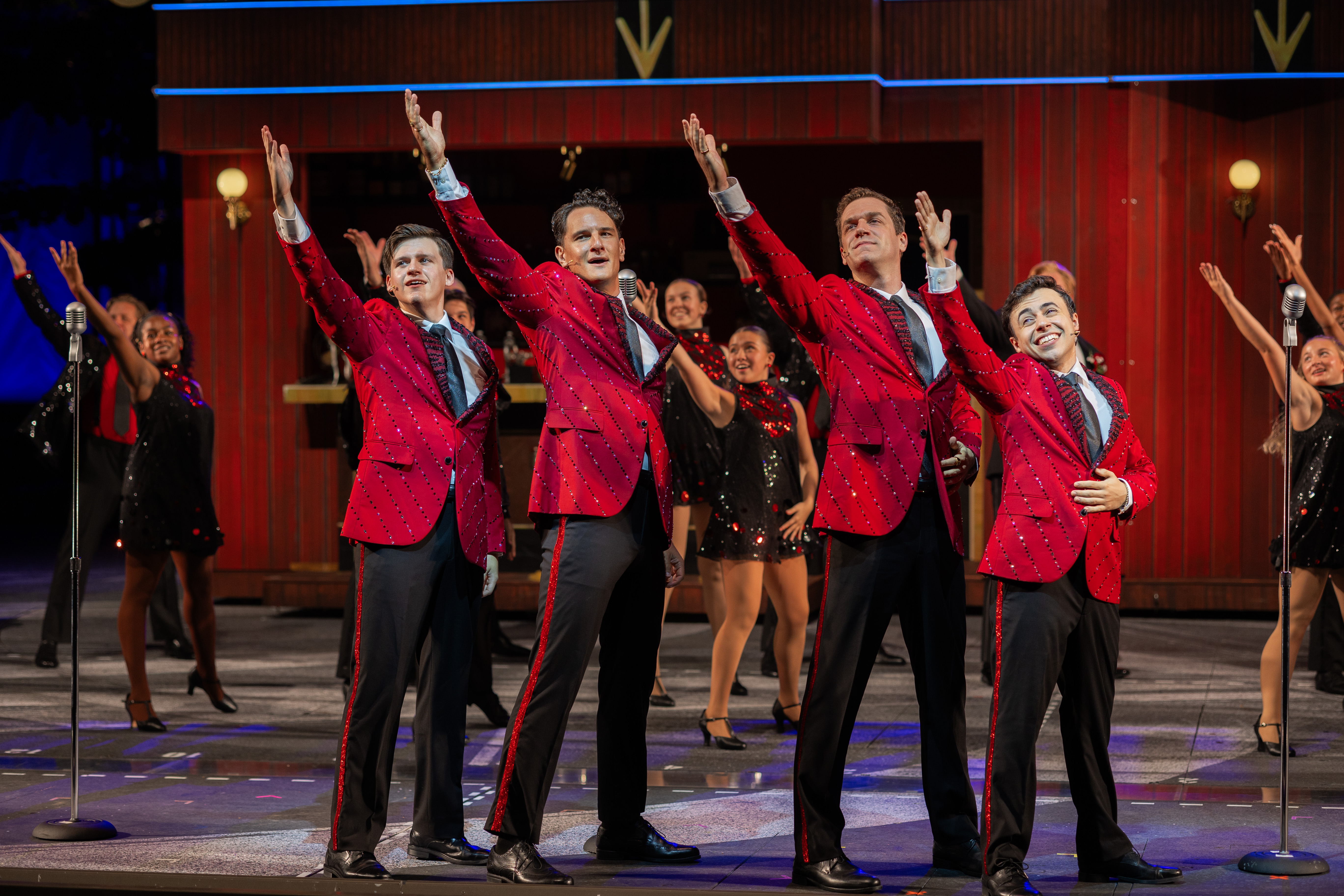 Four performers in matching red, sparkling jackets and black pants stand in front of a lively ensemble, raising one arm in a choreographed gesture on stage during a musical—an unforgettable night with exciting Ticket Savings available.