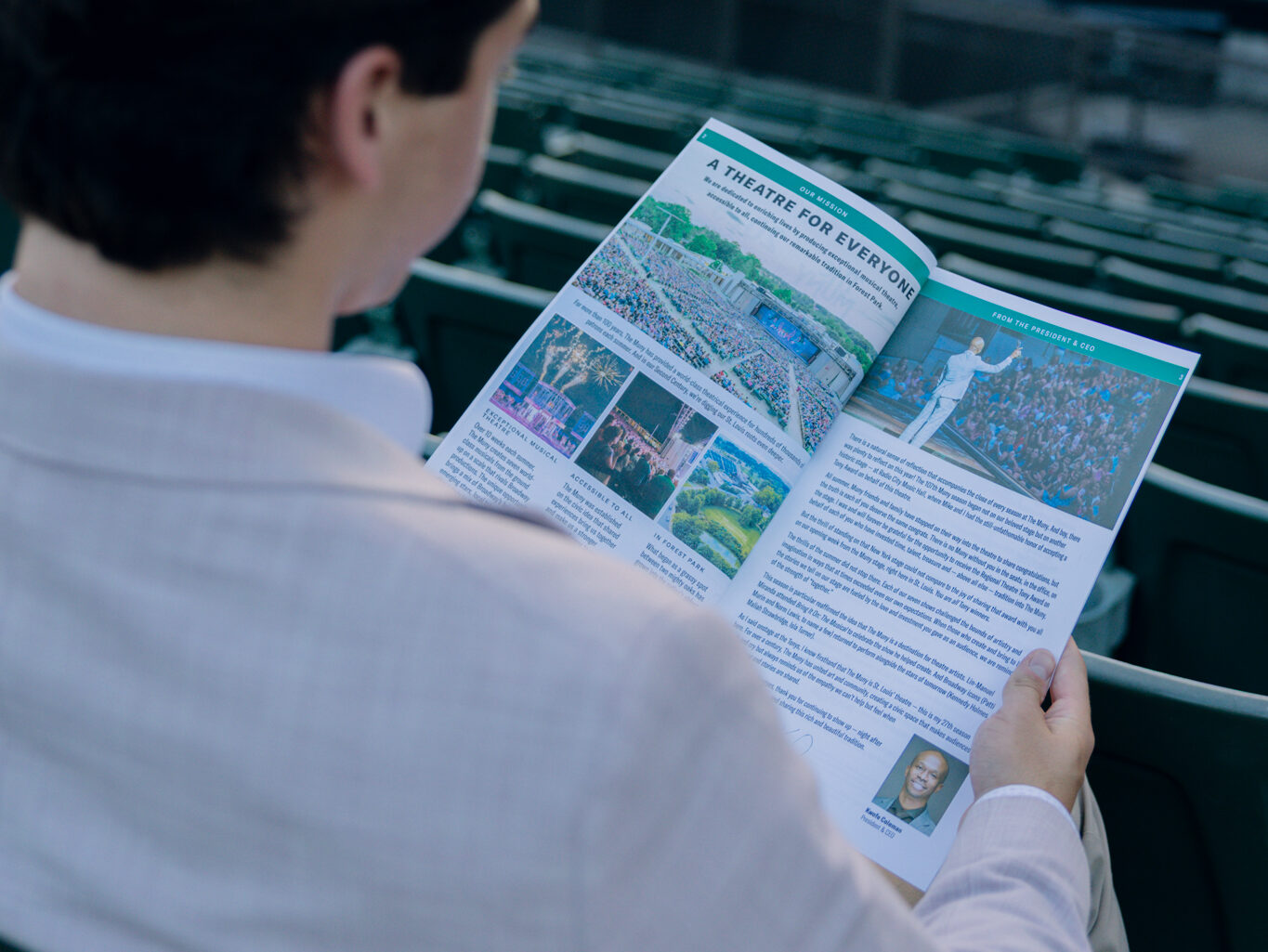 A person in a light-colored blazer sits in green theater seats, reading an accessible newsletter or brochure with photos and text. A large blurry JERSEY BOY sign is visible in the background.