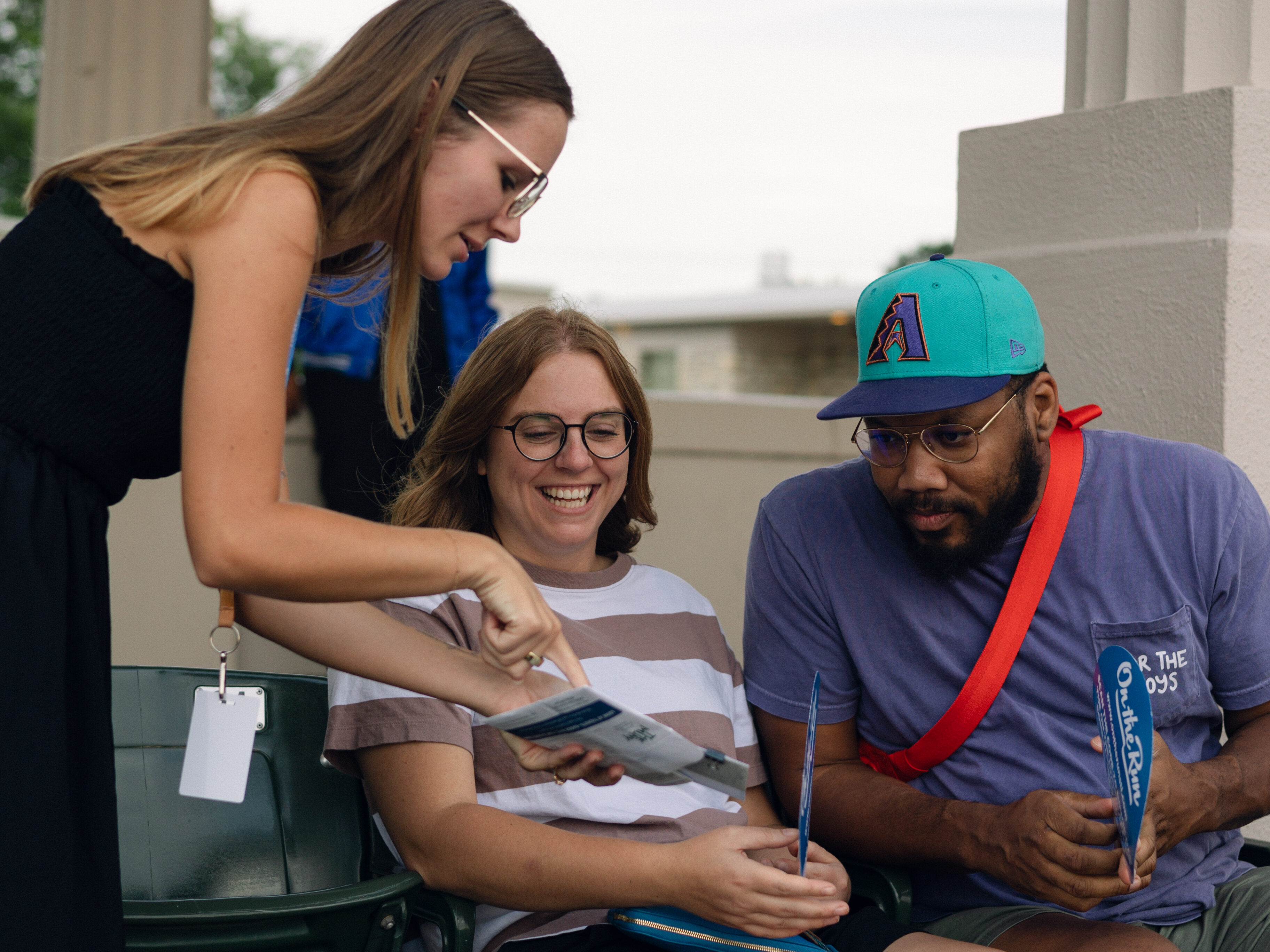 A woman stands and shows a magazine to a smiling seated woman and a man in a teal cap, discussing Ticket Savings in an outdoor stadium with green seats labeled 34 and 32.