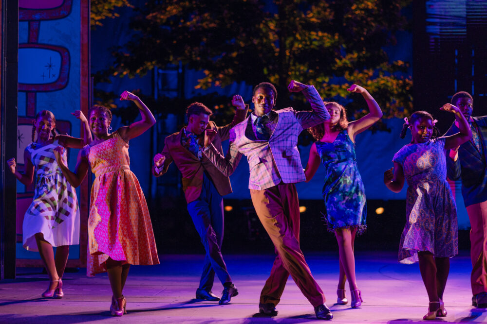 Muny Kids & Teens performers in colorful costumes dance energetically on stage at night, striking bold poses with bent arms under vibrant stage lighting. Trees and stage props add depth to the lively scene.