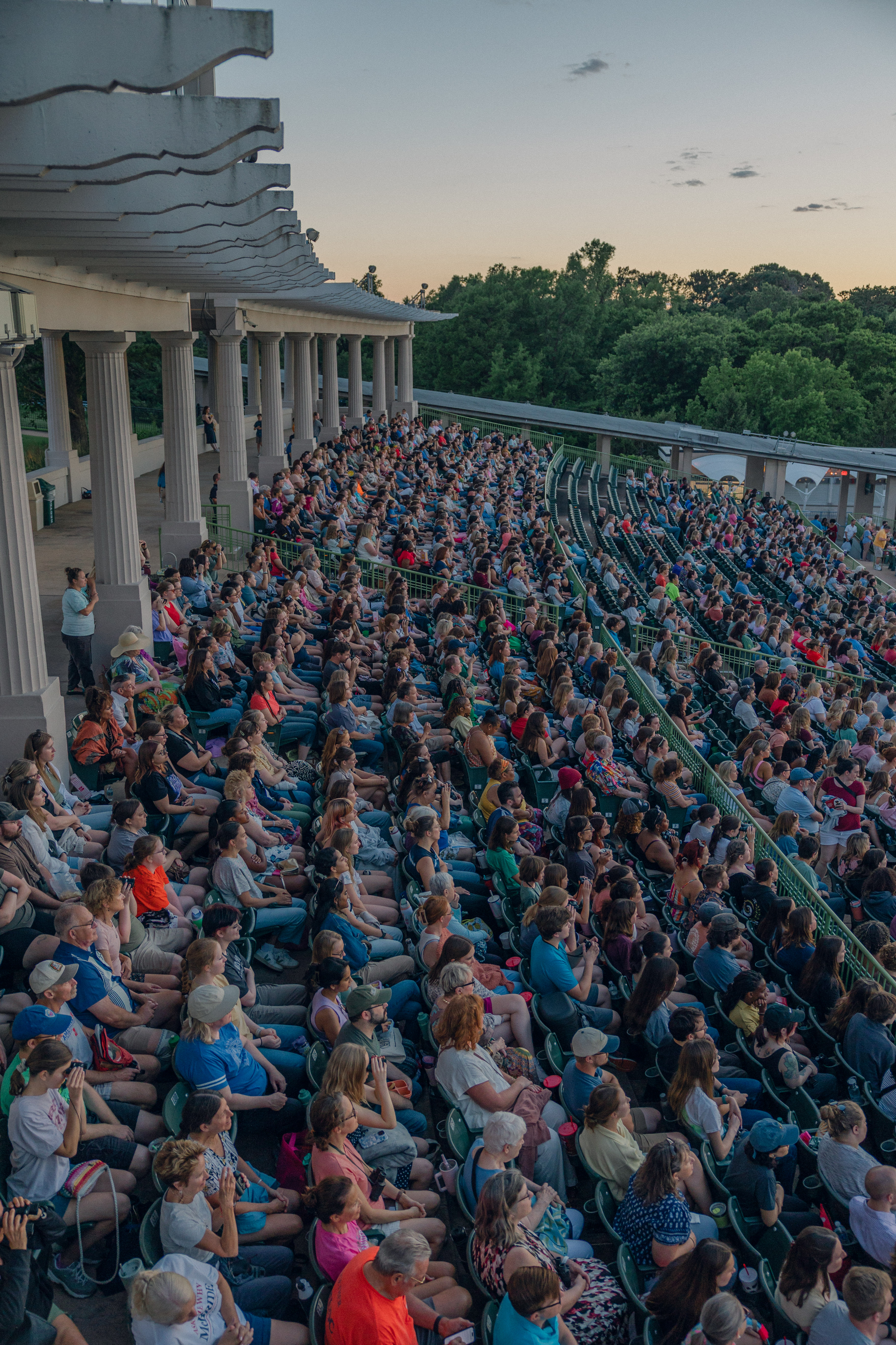 A large audience enjoys ticket savings as they sit in rows under an open-air pavilion at sunset, facing a stage out of view. The diverse crowd relaxes while trees frame the scenic background.