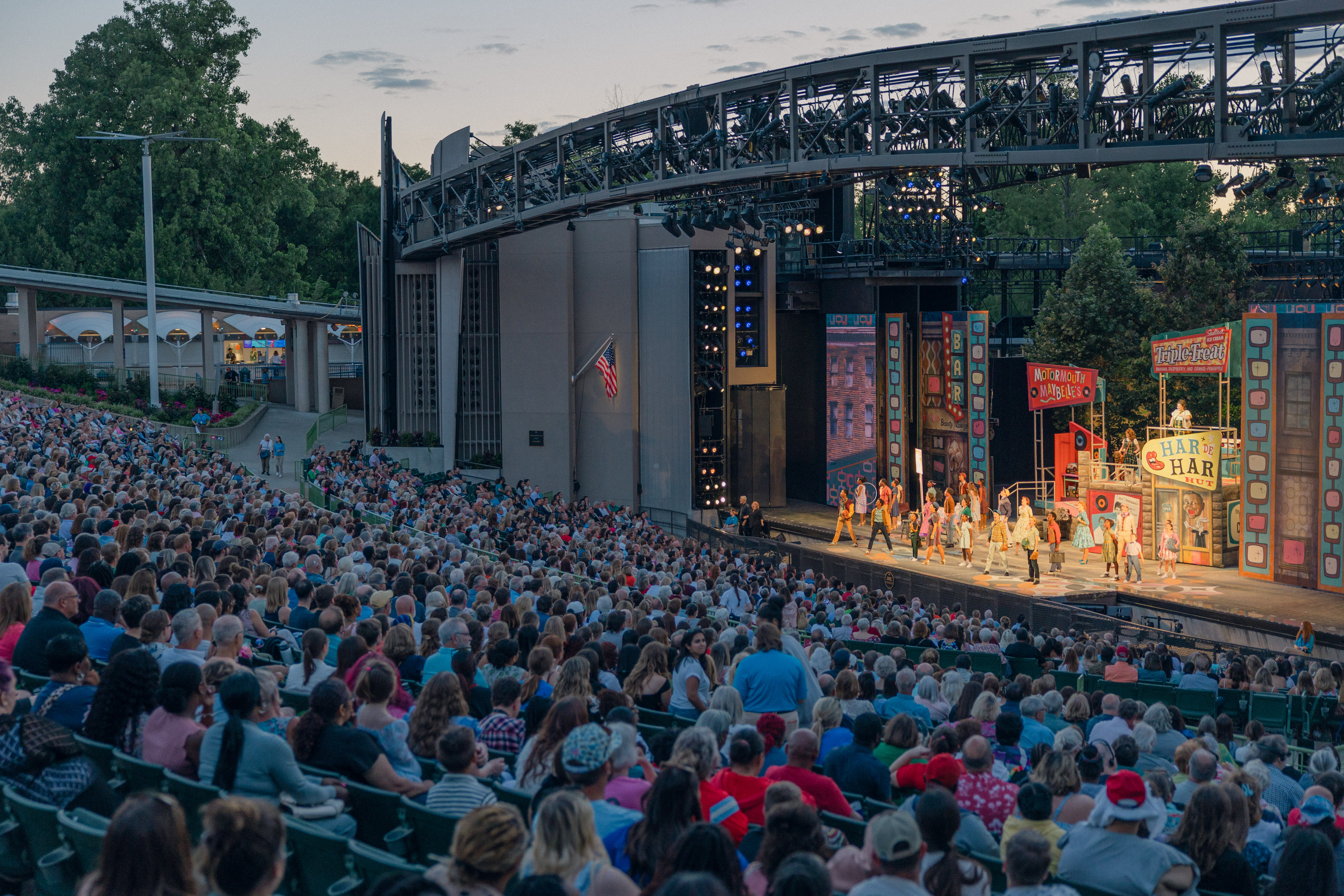 A large outdoor audience enjoys a colorful musical performance on an open-air stage at dusk, with bright lights, vibrant signs, and trees in the background—perfect for those seeking unforgettable moments and ticket savings.