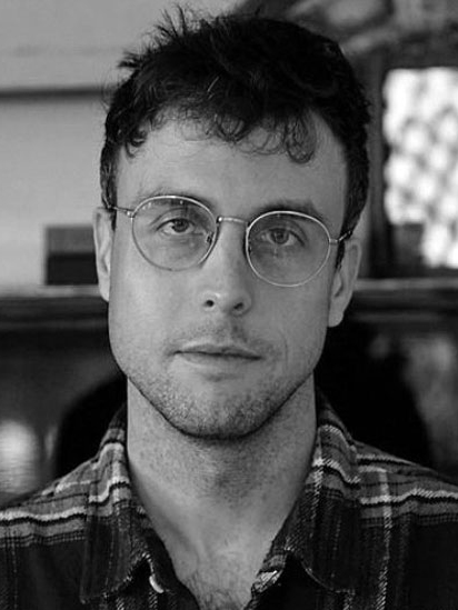 A man with short dark hair, wearing round glasses and a plaid shirt, looks at the camera with a neutral expression. The image is in black and white.
