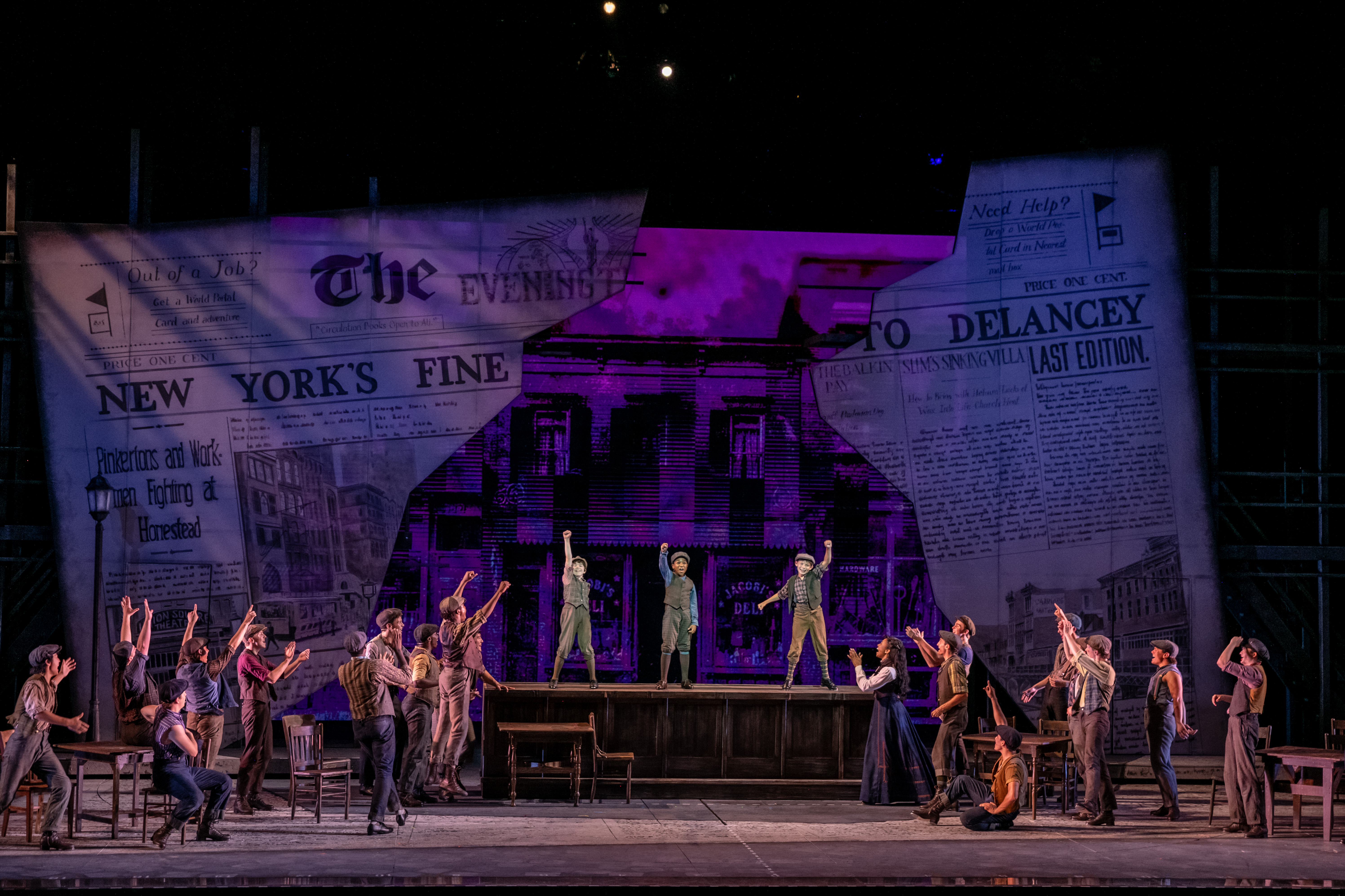 A stage performance features actors dressed as 19th-century newsboys, with large newspaper props as the backdrop and purple lighting highlighting the scene. Reminiscent of lessons learned in Education Classes, some actors stand on tables while others cheer with raised arms, bringing history to life through dynamic storytelling.