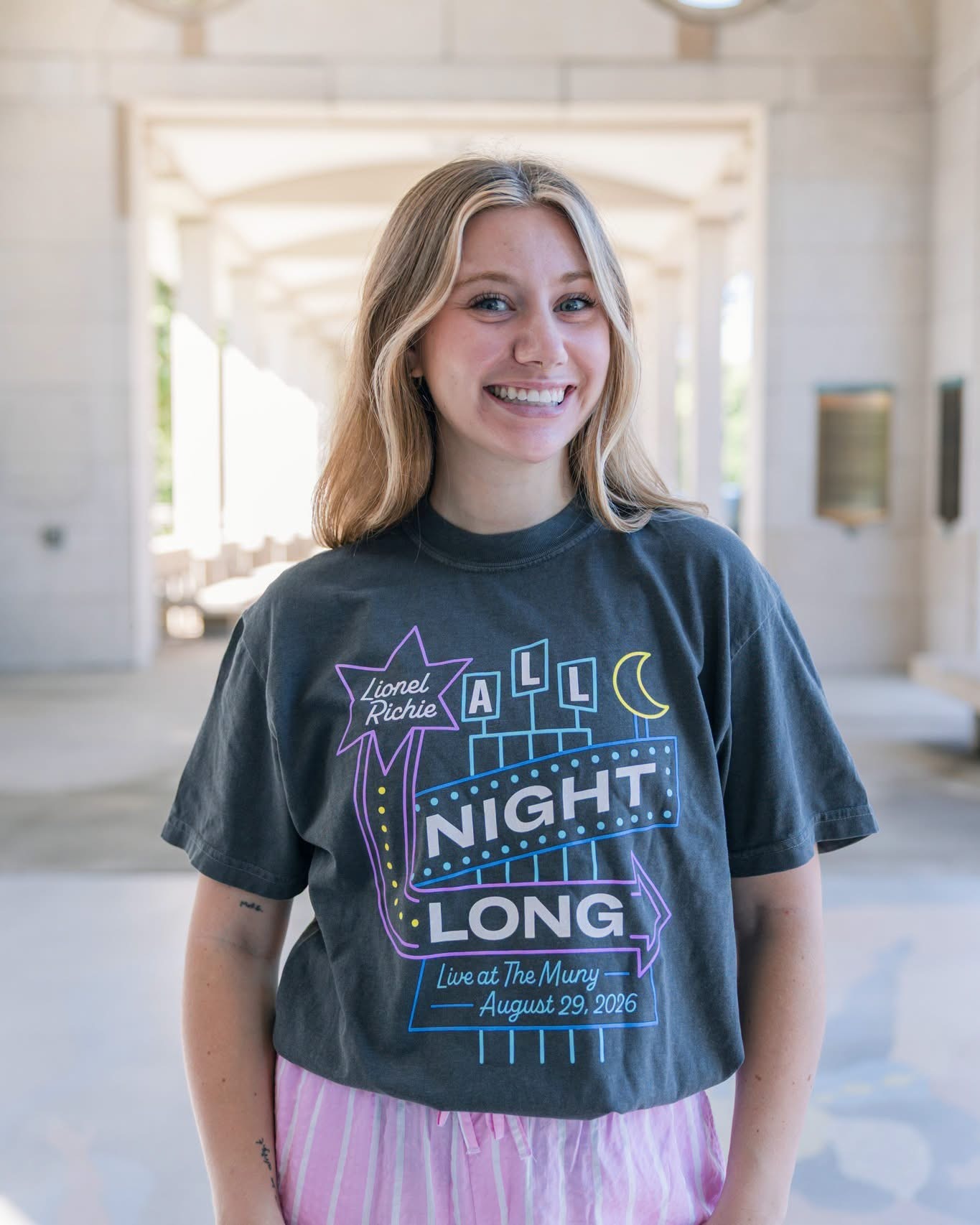 A woman with long blonde hair smiles as she stands in a sunlit corridor with columns, wearing a dark Meet Me In St. Louis Show Shirt.