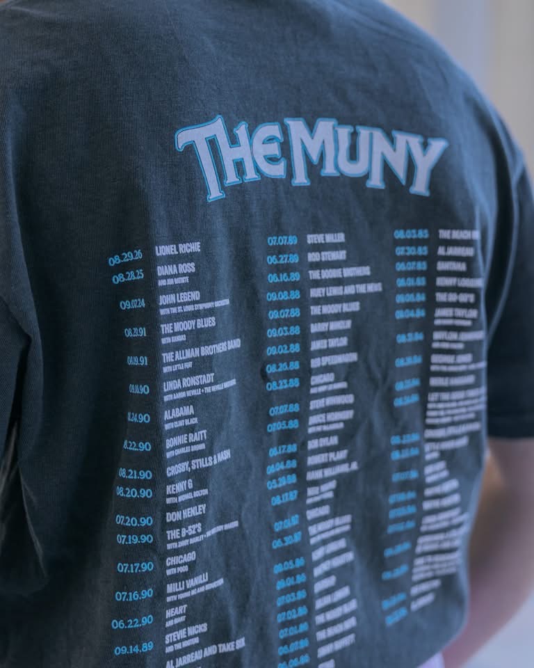 A person wearing the Meet Me In St. Louis Show Shirt, a black tee featuring The Muny and a list of artists and concert dates printed on the back in blue and white text.