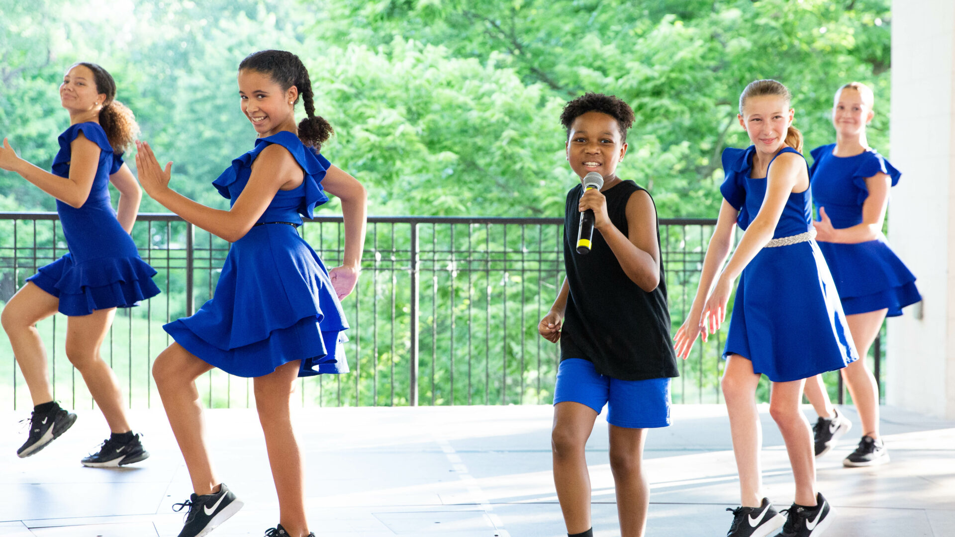 A boy singing into a microphone during an education performance as four girls in blue dresses dance behind him on an outdoor stage.